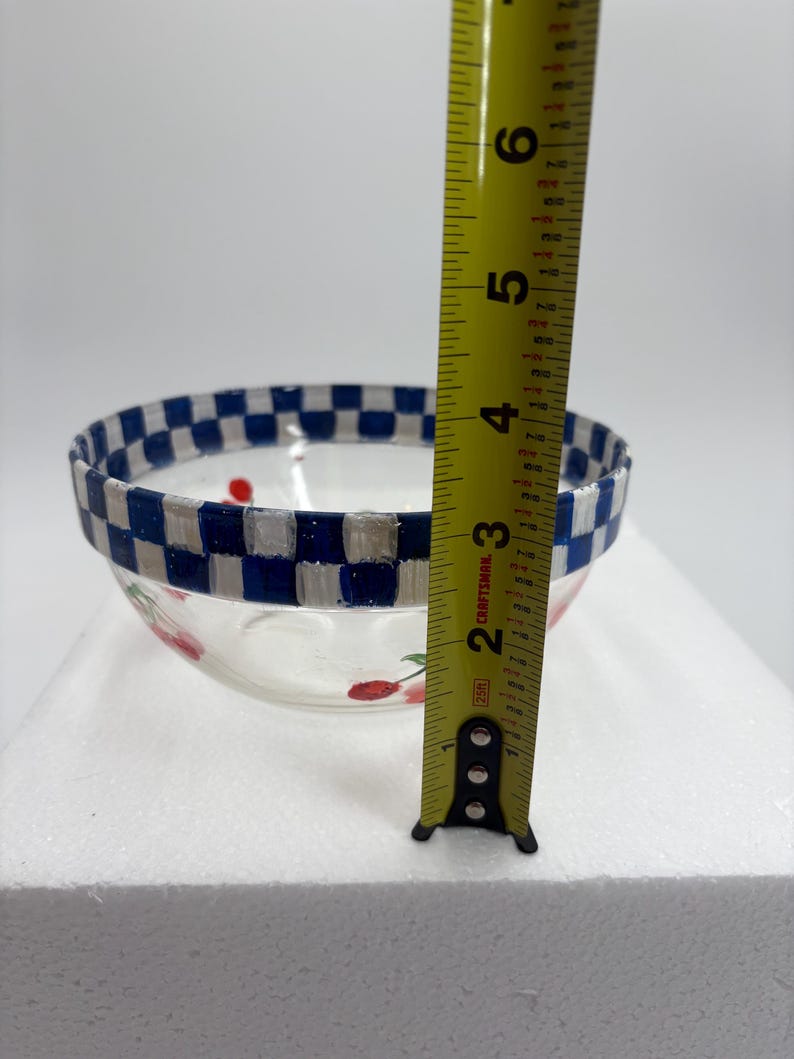May include: A clear glass bowl with a blue and white chequered rim and red cherry designs. The bowl is approximately 10 cm in height. The bowl is sitting on a white surface.
