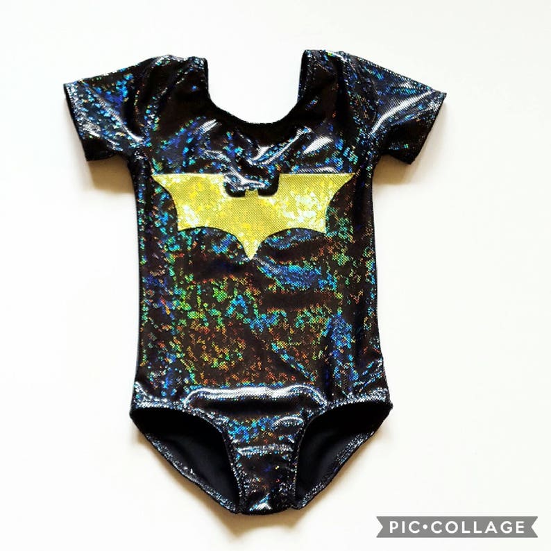 May include: Black and iridescent bodysuit with short sleeves and a yellow bat symbol on the front. The shiny fabric reflects multiple colors. The bodysuit is likely for a child or infant.