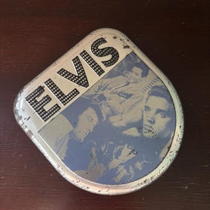 May include: A rounded square metal tin featuring the name ELVIS in bold black letters with a dotted pattern. The surface displays a blue-toned photographic collage of Elvis Presley playing a guitar and singing into a microphone with visible wear.