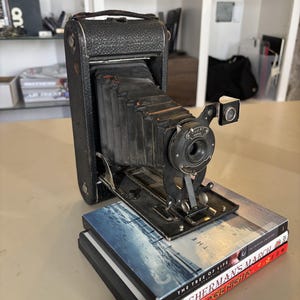 May include: A vintage black folding camera with a textured leather-like body and an extendable bellows mechanism. The lens assembly features a circular metal plate with the text Marvel. It is resting on a stack of three DVD cases.