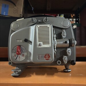 May include: A vintage grey Paillard Bolex 18-5 film projector featuring a prominent red speed control dial marked with R, 5, and 18. The front panel displays a white speaker grille, a lens assembly, and a label reading 18-5 Bolex Paillard.