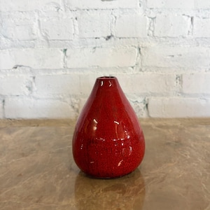 May include: A teardrop-shaped, red ceramic vase with a glossy finish. The vase has a narrow neck and a wide, rounded base. The surface has a speckled texture. The background is a white brick wall.
