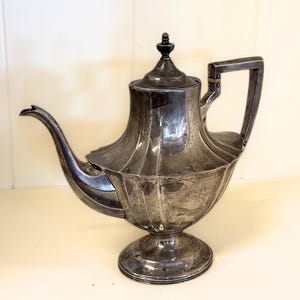 May include: A silver-toned metal teapot featuring a fluted, bulbous body, a curved spout, and a tall, angular handle. The lid is topped with a small, pointed finial. The surface displays a tarnished, weathered patina with visible vertical ridges.