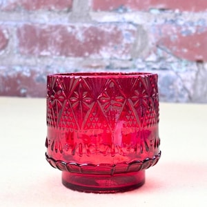 May include: A ruby red glass votive candle holder. The candle holder has a geometric pattern and a decorative base. The glass is transparent, allowing light to pass through. The candle holder is approximately 3 inches tall.