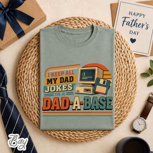 Op de afbeelding: Lichtgroen t-shirt met de tekst "I KEEP ALL MY DAD JOKES IN A DAD-A-BASE" en een retro computer graphic. Een Vaderdagkaart, een horloge en een geschenkdoos zijn ook zichtbaar.