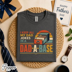 Op de afbeelding: Donkergrijs t-shirt met de tekst "I KEEP ALL MY DAD JOKES IN A DAD-A-BASE" in retro-ontwerp. De afbeelding bevat een archiefkast en een zonsondergang. Een Vaderdagkaart, een horloge en een geschenkdoos zijn ook zichtbaar.