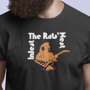 以下が含まれることがあります： 「The Rat'z Nest」の文字と、ギターを演奏する人のオレンジ色のイラストが描かれたグラフィックデザインの黒いTシャツ。デザインには「Infest」の文字も含まれています。サングラスをかけた人が着ています。