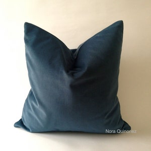 May include: A square, teal-colored velvet pillow. The pillow is a solid color and has a soft, plush appearance. The pillow is placed against a white background. The name "Nora Quinonez" is printed on the bottom right corner.