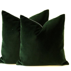 Pode incluir: Duas almofadas de veludo verde escuro sobre um fundo branco. As almofadas quadradas t&ecirc;m uma apar&ecirc;ncia macia e felpuda. A cor verde esmeralda profunda adiciona um toque de luxo e sofistica&ccedil;&atilde;o a qualquer ambiente.