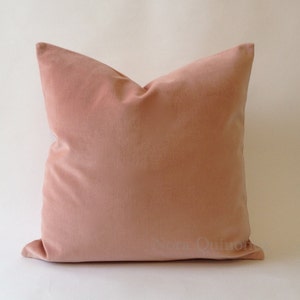 May include: A square, dusky pink velvet cushion. The cushion has a soft, plush appearance and is likely used for decorative purposes. The cushion is a solid colour and has a simple, elegant design. The text "Nora Quinon" is visible on the bottom right corner.