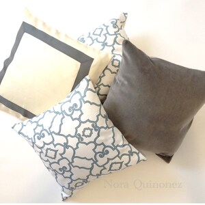 May include: Four decorative cushions in different designs. One is plain grey, another is cream-coloured with a grey border, and two have a white background with a blue geometric pattern. The cushions are arranged on a white surface.