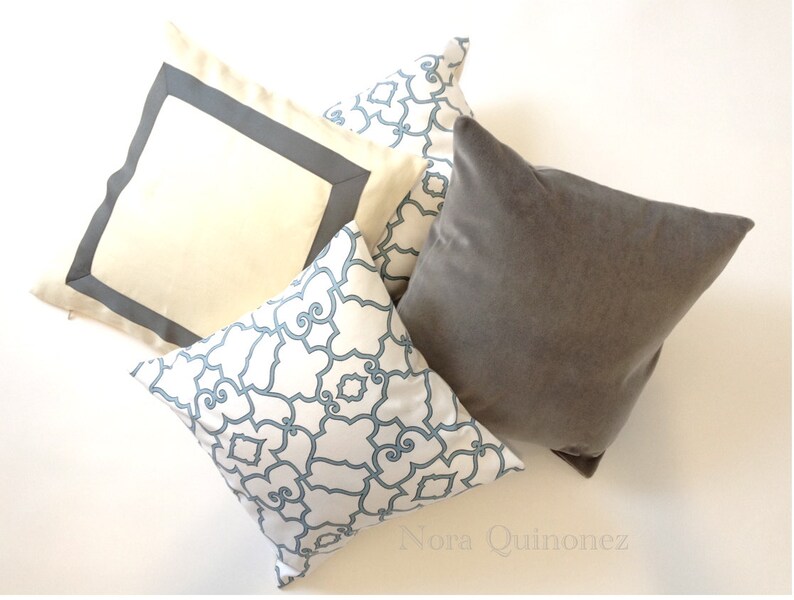 May include: Four decorative cushions in different designs. One is plain grey, another is cream-coloured with a grey border, and two have a white background with a blue geometric pattern. The cushions are arranged on a white surface.