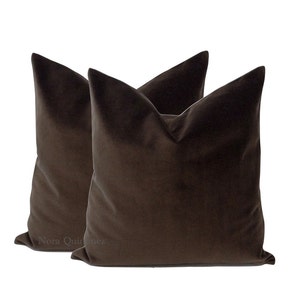 May include: Two brown velvet throw pillows. The pillows have a square shape with a pointed top. The fabric has a soft, plush appearance. The pillows are a solid, dark brown color.