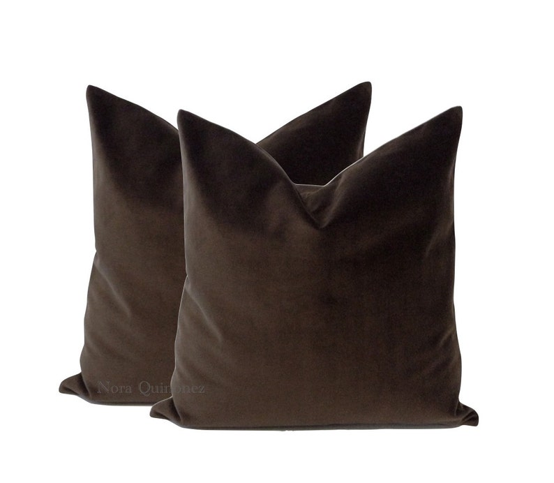 May include: Two brown velvet throw pillows. The pillows have a square shape with a pointed top. The fabric has a soft, plush appearance. The pillows are a solid, dark brown color.