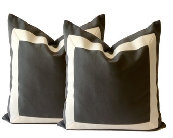 May include: Two square throw pillows. The pillows are a dark gray color with a wide, off-white border. The pillows are made of a soft fabric and are likely used for home decor. The pillows are placed on a white background.