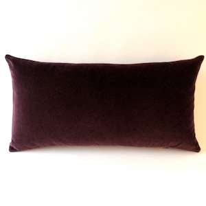 Puede incluir: Un cojín rectangular de terciopelo color ciruela. El cojín tiene un aspecto suave y afelpado y una forma ligeramente redondeada. El cojín está diseñado para uso decorativo en una cama o sofá.