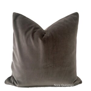 May include: A square, dark grey velvet cushion with a soft, plush appearance. The cushion has a classic shape and is likely used for decorative purposes on a bed or sofa. The cushion is against a white background.