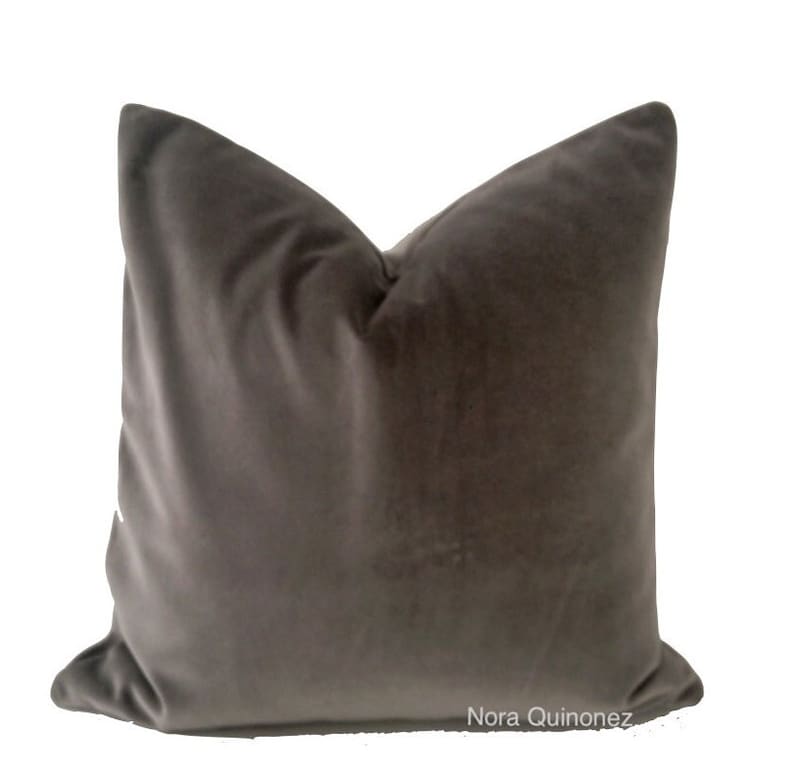 May include: A square, dark grey velvet cushion with a soft, plush appearance. The cushion has a classic shape and is likely used for decorative purposes on a bed or sofa. The cushion is against a white background.