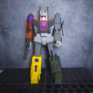 Puede incluir: Un juguete robot grande y colorido con un diseño detallado. El robot presenta un cuerpo gris, piernas amarillas y brazos marrones y grises. Tiene un cohete en un brazo y una pistola en el otro. La cabeza del robot tiene ojos rojos y un emblema púrpura.