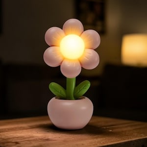 Könnte beinhalten: Eine rosa blütenförmige Lampe mit leuchtend gelbem Zentrum, grünem Stiel und Blättern, in einem passenden rosa Topf. Die Lampe steht auf einem Holztisch und schafft eine warme, einladende Atmosphäre. Die Blumenlampe ist eine dekorative Leuchte.