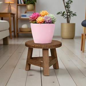 May include: A pink flower pot filled with colorful artificial succulents sits on a small, round, wooden stool. The stool has a rustic, weathered appearance. The background includes a sofa, a lamp, and other decorative items, suggesting an interior home decor theme.