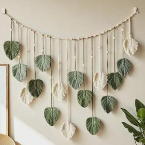 May include: A macrame wall hanging featuring leaf-shaped designs in shades of green and cream. The leaves are suspended from a wooden dowel with a string and bead design. The wall decor adds a natural, bohemian touch to any room.
