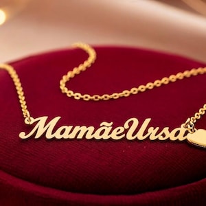 Op de afbeelding: Een goudkleurige ketting met de naam "Mamãe Ursa" in cursief schrift. Een kleine hartvormige bedel hangt eraan. De ketting ligt op een rode fluwelen ondergrond.
