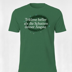 May include: Green t-shirt with the German phrase "Träume heller als die Schatten deiner Ängste" printed in white. The text is inside a dark oval with small star accents. The shirt is a solid green colour.