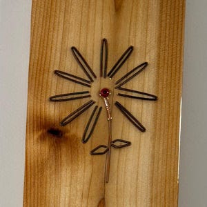 May include: Wooden plaque with a handcrafted flower design. The flower features dark wire petals, a copper stem, and a red bead center. The wood has a natural grain and a warm, light brown color.