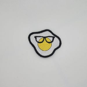Op de afbeelding: Geborduurde patch met een gebakken ei-ontwerp met een zwart montuur. Het ei heeft een gele dooier, een wit eiwit en een zwarte omtrek. De patch is waarschijnlijk voor kleding of accessoires.
