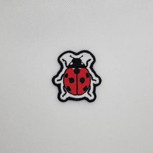 May include: Embroidered ladybird patch featuring a red body with black spots, a black head, and a white border. The patch is set against a white fabric.