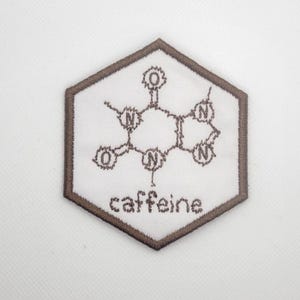 Peut inclure: Écusson hexagonal blanc avec une bordure marron. L'écusson présente un motif brodé de molécule de caféine et le mot "caffeine" dans une police simple. Le motif est en fil marron.