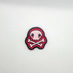 May include: Embroidered patch featuring a cartoon skull with crossbones. The skull is pink with black eyes and a red outline. The crossbones are white, and the patch has a black border. This patch is suitable for jackets, bags, or other fabric items.