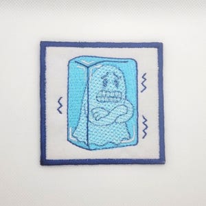May include: Embroidered patch featuring a light blue ghost-like figure encased in a blue ice block. The figure has a distressed expression, with zig-zag lines on the sides. The patch has a white background with a dark blue border.
