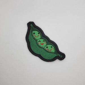 Puede incluir: Parche bordado con una vaina de guisantes verde con tres guisantes sonrientes. La vaina de guisantes tiene un borde negro. Los guisantes tienen caras sonrientes con bocas rojas y ojos negros. Ideal para ropa o accesorios.