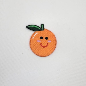May include: A circular orange embroidered patch featuring a cartoon face with black eyes, a red curved mouth, and pink cheeks. The orange has a single green leaf attached to the top and is outlined with a thick orange thread border.