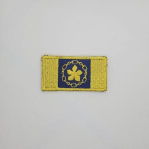 May include: Embroidered patch with a yellow background and a blue square in the center. The blue square features a yellow flower design surrounded by a chain. The patch has a rectangular shape.