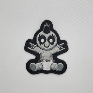 Peut inclure: Écusson brodé représentant un bébé stylisé avec une crête, du maquillage noir pour les yeux et une couche. Le bébé fait un signe de la main rock and roll. L'écusson a une bordure noire et est en nuances de gris et de blanc.