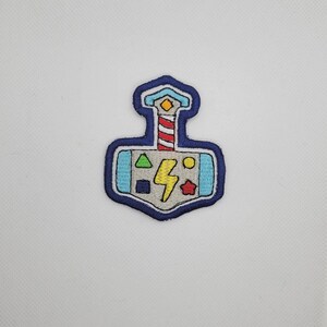 May include: Embroidered patch in the shape of a hammer, featuring a lightning bolt, geometric shapes, and a lighthouse design. The patch has a blue border and is set against a white background. This patch is suitable for clothing or accessories.
