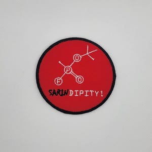 Puede incluir: Un parche redondo rojo con borde negro. Presenta una estructura química blanca estilizada y la palabra "SARIN" tachada, con "DIPITY!" debajo. El parche es probablemente un artículo bordado.