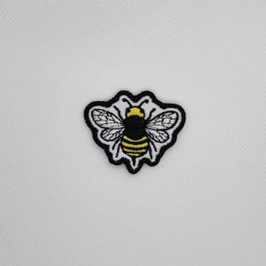 May include: Embroidered bee patch with a black outline and a white background. The bee has yellow and black stripes on its abdomen and yellow on its head. The wings are white with black detailing.