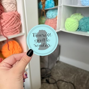 May include: A round, light blue sticker with the text "Dearest gentle reader" in black script. The sticker features a gray silhouette of a woman's profile within a black border. The background includes colorful yarn balls.