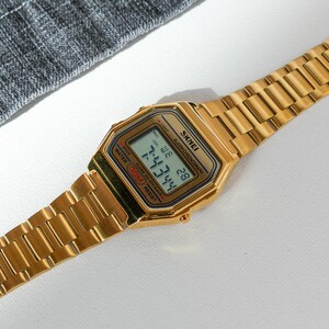May include: A gold-toned digital watch with a rectangular face and matching band. The watch face displays the time, date, and other functions. The watch has the brand name "SKMEI" printed on the face.