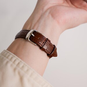 May include: A brown leather watch band with a silver buckle is fastened on a wrist. The band has a textured, crocodile-like pattern. The watch band is a rich, dark brown color.