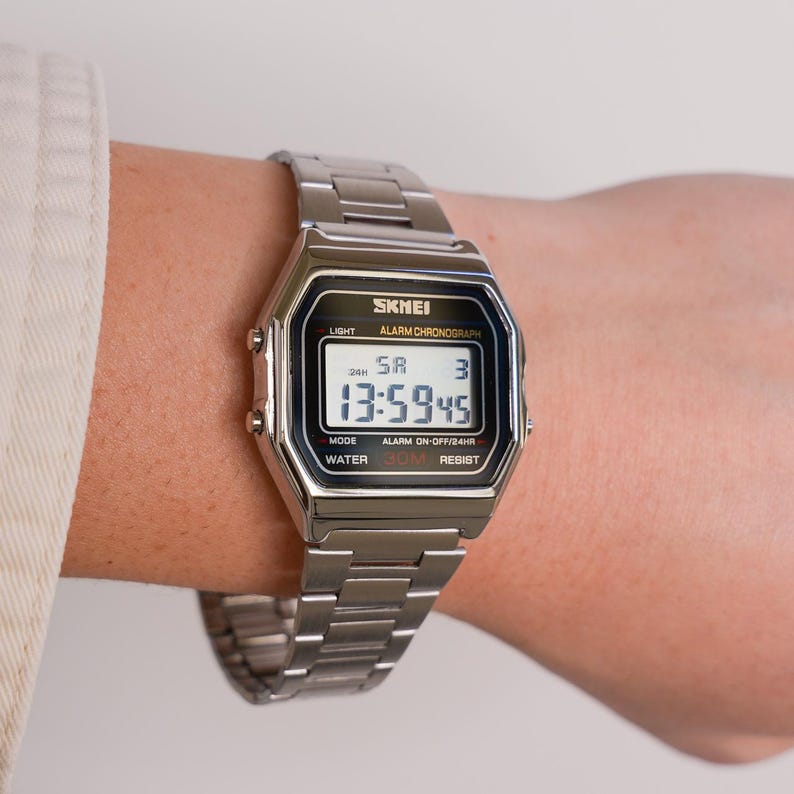Puede incluir: Un reloj digital plateado con una esfera rectangular y una correa de metal a juego. La esfera del reloj muestra la hora, el d&iacute;a y la fecha. El reloj tiene la marca "SKMEI" y otros textos como "LIGHT", "ALARM CHRONOGRAPH", "WATER 30M RESIST".