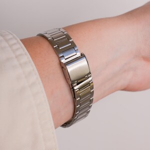Puede incluir: Una correa de reloj de metal plateado est&aacute; abrochada alrededor de una mu&ntilde;eca. La correa est&aacute; hecha de eslabones rectangulares y tiene un cierre seguro. La correa del reloj se muestra sobre un fondo neutro.