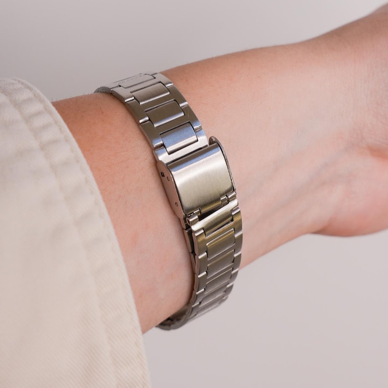 Puede incluir: Una correa de reloj de metal plateado est&aacute; abrochada alrededor de una mu&ntilde;eca. La correa est&aacute; hecha de eslabones rectangulares y tiene un cierre seguro. La correa del reloj se muestra sobre un fondo neutro.