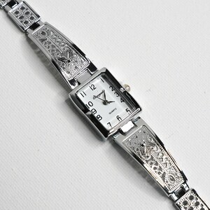 May include: Silver-toned quartz watch with a rectangular white face and black numerals. The watch band is intricately designed with a decorative pattern. The watch face reads "Quartz" and "Orogold".