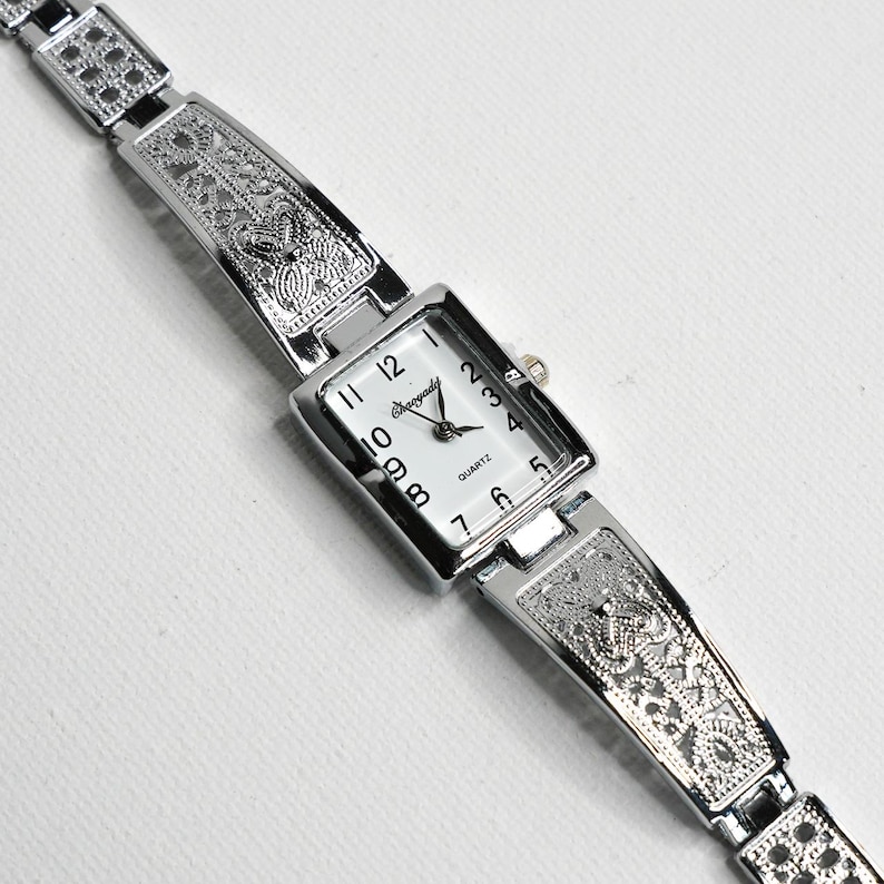 May include: Silver-toned quartz watch with a rectangular white face and black numerals. The watch band is intricately designed with a decorative pattern. The watch face reads "Quartz" and "Orogold".