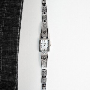 May include: A silver-toned rectangular watch with a matching bracelet. The watch face displays the time with black numerals and hands. The bracelet and watch casing feature decorative patterns.